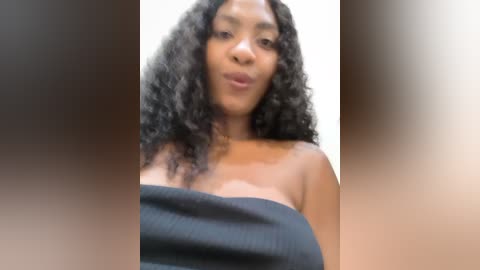 Video of a Black woman with a medium brown complexion and curly black hair, wearing a black strapless top, making a pouty face, with blurred background.