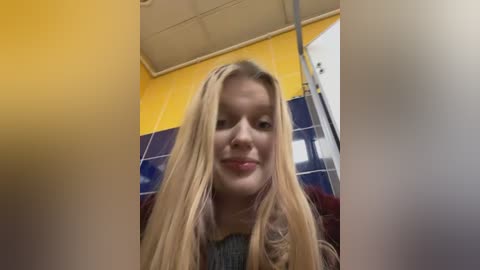 A video of a young woman with long, straight blonde hair, fair skin, and a slight smile. She wears a gray sweater. The background shows a yellow wall, blue tiles, and a showerhead.