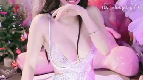 Video of a smiling Asian woman in a revealing white lace bra, seated on a bed with a Christmas tree in the background.
