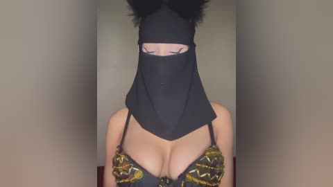 Media: Video of a woman with a black face mask, black headpiece resembling cat ears, and a bra with gold and black sequins, revealing ample cleavage. Background is a plain beige wall.