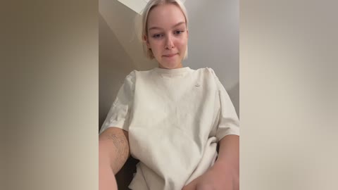 Video of a young Caucasian woman with short, platinum blonde hair, wearing a loose-fitting white T-shirt, standing indoors against beige walls.