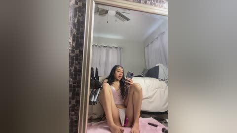 Video of a young woman with long black hair, wearing pink lingerie, sitting on a pink blanket in front of a mirror in a modestly decorated bedroom.