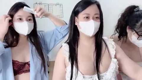 Video of three East Asian women in casual attire, wearing face masks, with one adjusting a mask on another's head. Background includes a calendar and white walls.