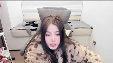 Video of a young woman with long, straight black hair, pale skin, and light makeup, sitting on a beige couch with a leopard-print blanket, in a modern, minimalistic living room with white walls and a window.