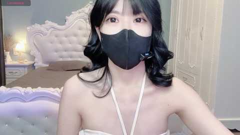 Video of an Asian woman with shoulder-length black hair, wearing a black face mask and a white strapless dress, sitting in a plush, white tufted bed with a nightstand and lamp in a light blue room.