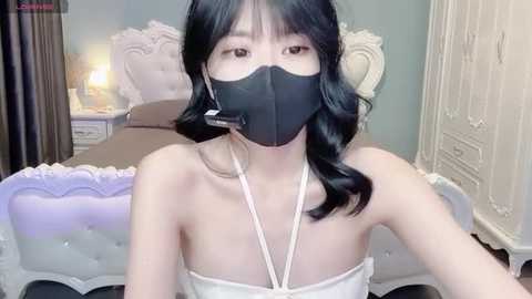 Video of an East Asian woman with long black hair, wearing a black face mask, white strapless top, and sitting on a pastel purple tufted bed with white headboard.