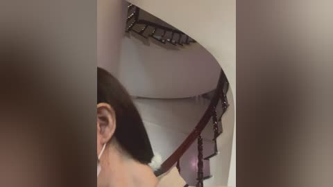 Video of a woman with short, dark hair, wearing white headphones, climbing a spiral staircase with a metal railing, taken from a low angle, blurred background.