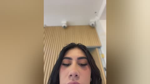 Video of a woman with long black hair, wearing a dark dress, standing in a beige hallway with wooden slats on the floor and white walls. Her eyes are closed, and she has full makeup.