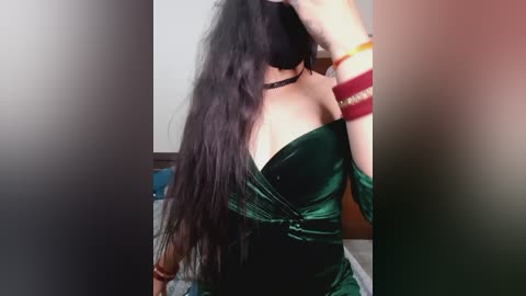 Video of a woman with long, wavy black hair, wearing a green velvet off-shoulder dress, multiple bangles on her wrist, and a black choker. Background is blurred.