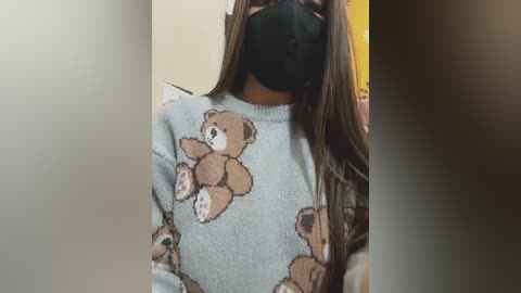 A video of a person with long brown hair, wearing a light blue sweater with bear patterns, black mask, and black face shield, standing indoors with blurred background.