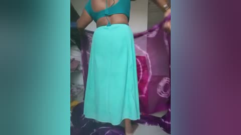 Video of a woman in a teal top and skirt, holding a pink and purple inflatable pool toy, standing in a cluttered room with white walls and scattered objects.