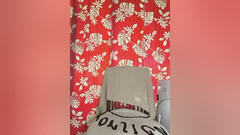 Video of a child's crib with a white crib sheet featuring black lettering, surrounded by a vibrant red curtain adorned with white hibiscus flowers.