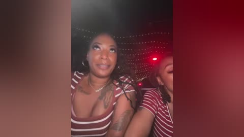 Video of a smiling Black woman with dark skin and medium-length black hair, wearing a low-cut red and white striped shirt, seated in a dimly lit bar with festive string lights and red glow.