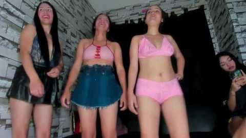 Video of four women in skimpy outfits: two in pink bikinis, one in a leather skirt, and one in a denim skirt, in a dimly lit room with brick walls.