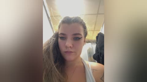 A video of a young woman with light skin and long, wavy, blonde hair, wearing a white tank top, touching her hair in a bathroom with a tiled ceiling.