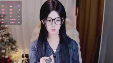 A video of a young woman with long black hair, wearing black-rimmed glasses, and a dark blue blouse, standing in a softly lit room with a Christmas tree in the background.