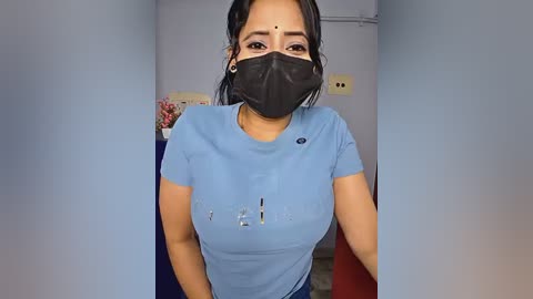 Video of a young woman with medium skin tone, wearing a light blue t-shirt, black mask, and earrings. Background shows a plain room with a red table and flowers.