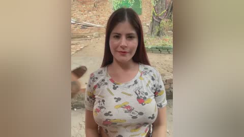 A video of a young, light-skinned woman with long, dark brown hair, wearing a tight, white T-shirt with colorful Minnie Mouse designs, standing on a dirt path in a rural setting with a brick wall and greenery in the background.
