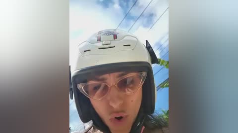 Video of a woman wearing a white helmet, clear safety glasses, and a pink shirt, seen from a low angle. She has a serious expression. Background shows a bright blue sky and green plants.
