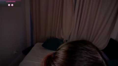 Video of a dimly-lit bedroom with a person lying face down on a bed, partially covered by a beige sheet. A small, dark object is visible on the pillow.