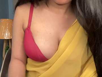 Video of a woman with long dark hair, wearing a yellow sari draped over a red bra, with a beige lamp and blue wall in the background.