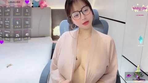 Video of an East Asian woman with fair skin, wearing glasses, a light pink robe, and a revealing neckline, sitting on a blue chair in a modern room.