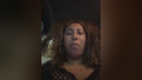 A dimly lit video shows a woman with curly hair and closed eyes, wearing a black lace top, sitting in a dark room.