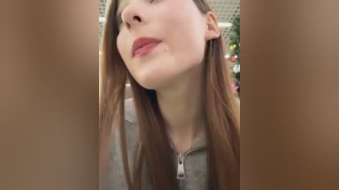 Video of a young Caucasian woman with long brown hair, light skin, and red lips, wearing a grey hoodie, looking up with a slight smile, blurred background.