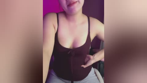 A video of a light-skinned woman with a small bust, wearing a black tank top and grey sweatpants, sitting with her hands on her hips, smiling. The background is blurred, suggesting a dimly lit room.