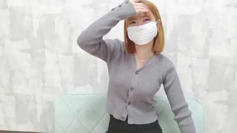 Video of a young Asian woman with straight shoulder-length blonde hair, wearing a white surgical mask, grey cardigan, and black pants, wiping her forehead, in a light blue upholstered chair against a textured, light-colored wall.