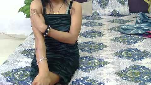 Video of a woman with medium-dark skin, wearing a black satin dress, sitting on a bed with intricate blue and white paisley-patterned bedding, leaning against a white wall.