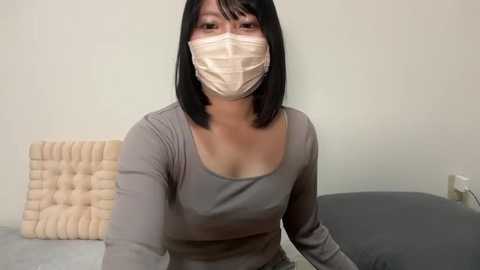 Video of a woman with straight black hair, wearing a gray long-sleeve top and beige face mask, sitting in a chair with a beige cushion, in a simple, neutral-toned room.