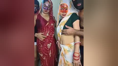 Video: Two women in vibrant Indian saris with elaborate makeup, red and green respectively, smiling widely. They are flanked by partially visible men. Background is indistinct.