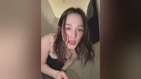 Video of a young, pale-skinned woman with wavy brown hair, wearing a white lace bra and black top, leaning forward in a dimly lit room.