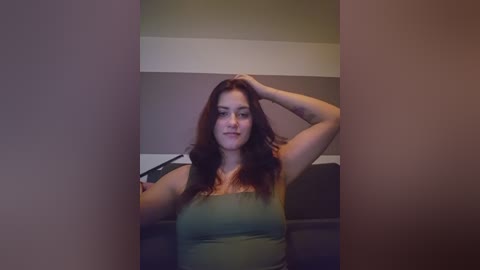 Video of a plus-sized woman with long, dark hair, wearing a green tank top, sitting on a dark couch in a dimly lit room.