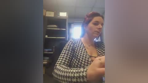 Video of a woman with dark hair, wearing a houndstooth-patterned cardigan, standing in a dimly lit office with gray carpeting and bookshelves in the background.