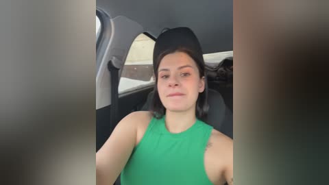 Video of a young woman with shoulder-length black hair, wearing a green tank top, sitting in a car with dark upholstery. The image is slightly blurry and has a grainy texture.