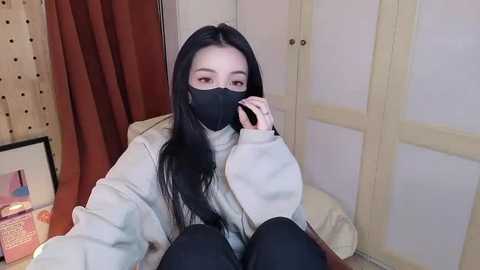 Video of an Asian woman with long black hair, wearing a gray sweater, black mask, and black pants, sitting indoors, looking contemplative. Background includes beige doors and a framed picture on a table.