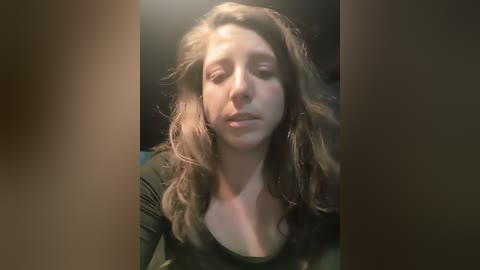 Video of a Caucasian woman with wavy, light brown hair, wearing a black top, sitting with a pensive expression, lit by soft, ambient light.