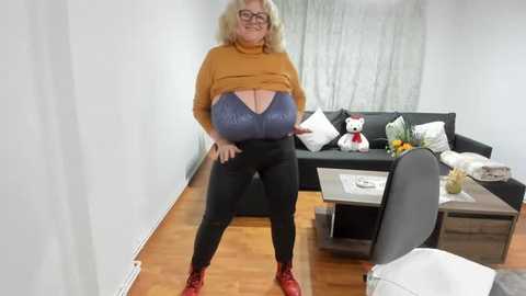 Video of a plus-size woman with large breasts, wearing a brown turtleneck, blue sports bra, black pants, and red shoes, standing in a modern living room with wooden floors, a black sofa, and a teddy bear.