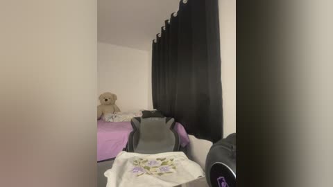 Video of a small, sparsely furnished room with white walls, a beige teddy bear, two unmade beds with pink and white sheets, a black curtain, and a black gaming chair.