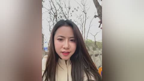 A video of a young Asian woman with long, dark hair and fair skin, wearing a beige coat, standing outdoors in a park with bare trees and a blurred background.