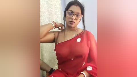 Video of a South Asian woman with medium skin tone, wearing a sheer red saree, exposing her bra, glasses, and a gold necklace.