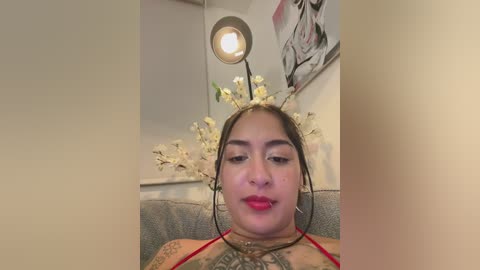 A video of a woman with light skin, dark hair, and tattoos, wearing red lipstick, sitting on a grey couch, adorned with floral headpiece, in a modern, minimalist room with a round lamp and abstract art.