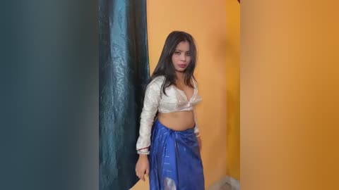 Video of a young woman with long, straight black hair, wearing a white lace bra and a blue saree, standing against vibrant orange and teal walls.