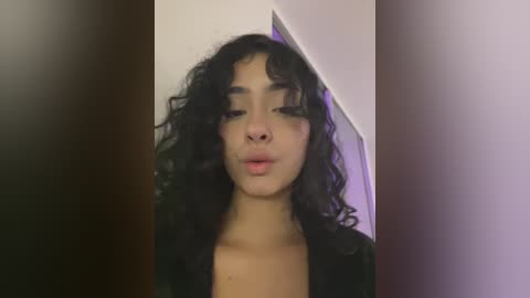 A video of a young woman with curly, dark hair and light skin, wearing a black top, blowing a kiss against a white wall with a purple shadow.