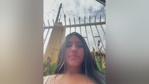 Video of a young woman with long dark hair, fair skin, and light makeup, standing near a tall, partially broken fence under a cloudy sky.