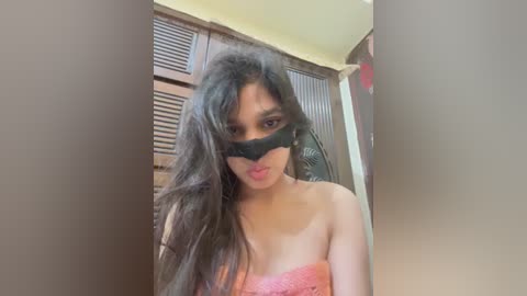 A video of a young Asian woman with long black hair and fair skin, partially covered by a black mask, wearing a pink strapless top. She stands in a dimly lit room with wooden shutters and a yellow wall.