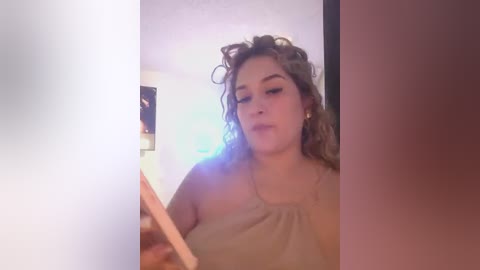 A candid video of a young woman with curly brown hair, fair skin, and a light beige tank top, standing in a dimly lit room with a white wall and a blurry painting.