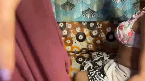 A video of a small child in a colorful, patterned room with a teal curtain, a brown and white rug, and a maroon garment. The child wears a floral-patterned top.